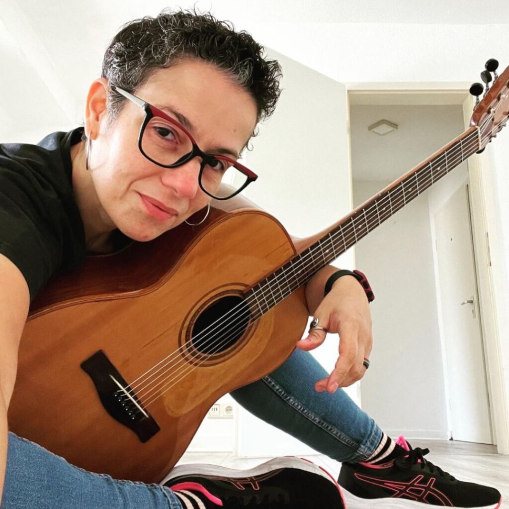 Aline Sardao sitzt mit einer Gitarre und blickt in die Kamera